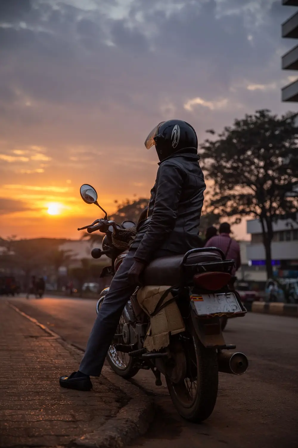 A boda boda rider pausing on a Kampala street at sunset